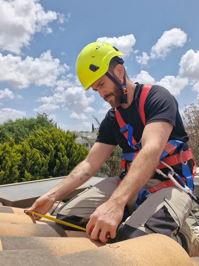עידו עם קסדה ורתמת בטיחות מודד רעף בסרט מדידה על גג רעפים מול שמיים כחולים